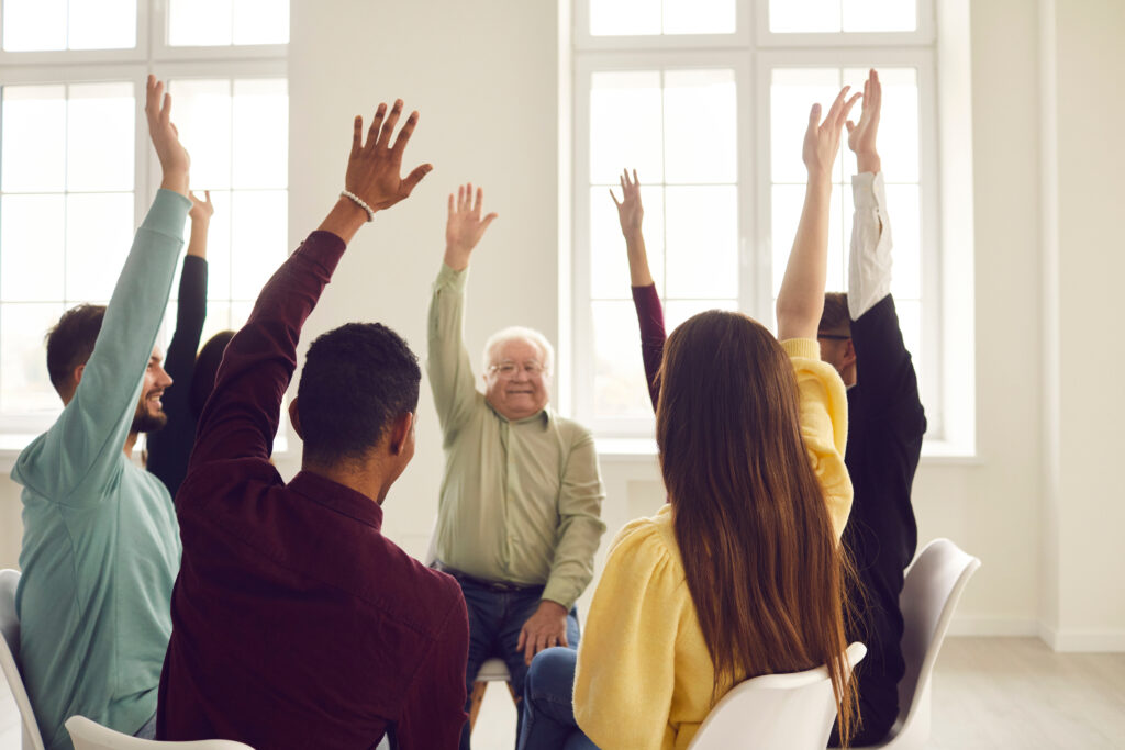 Group of happy diverse young and mature people in casual clothes sitting on chairs in circle and raising hands, approving of good idea and voting for useful suggestion in community club meeting